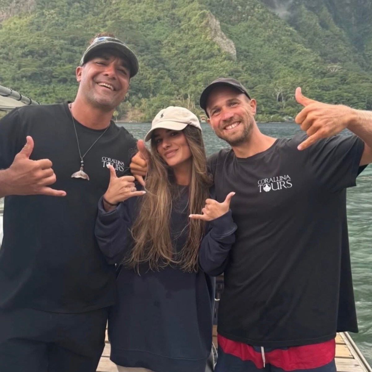 Three people posing on a boat with mountains in the background, making 'shaka' hand signs.