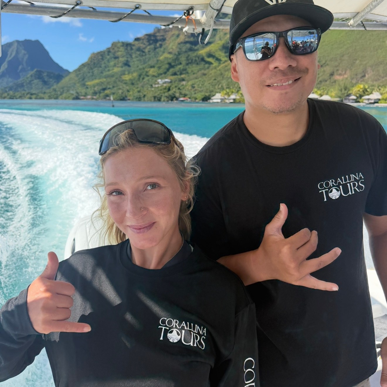 Two people on a boat, posing with shaka signs, mountains and water in the background.