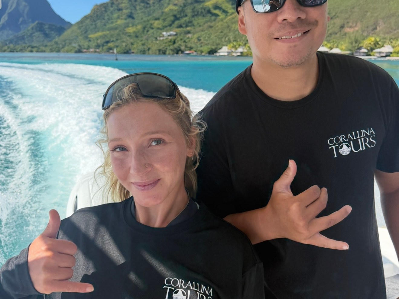 Two people on a boat, posing with shaka signs, mountains and water in the background.
