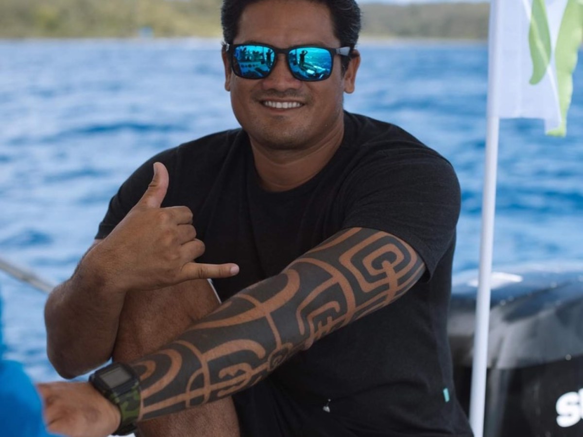 Man with tattooed arm on a boat, wearing sunglasses and a black shirt, making a shaka sign.