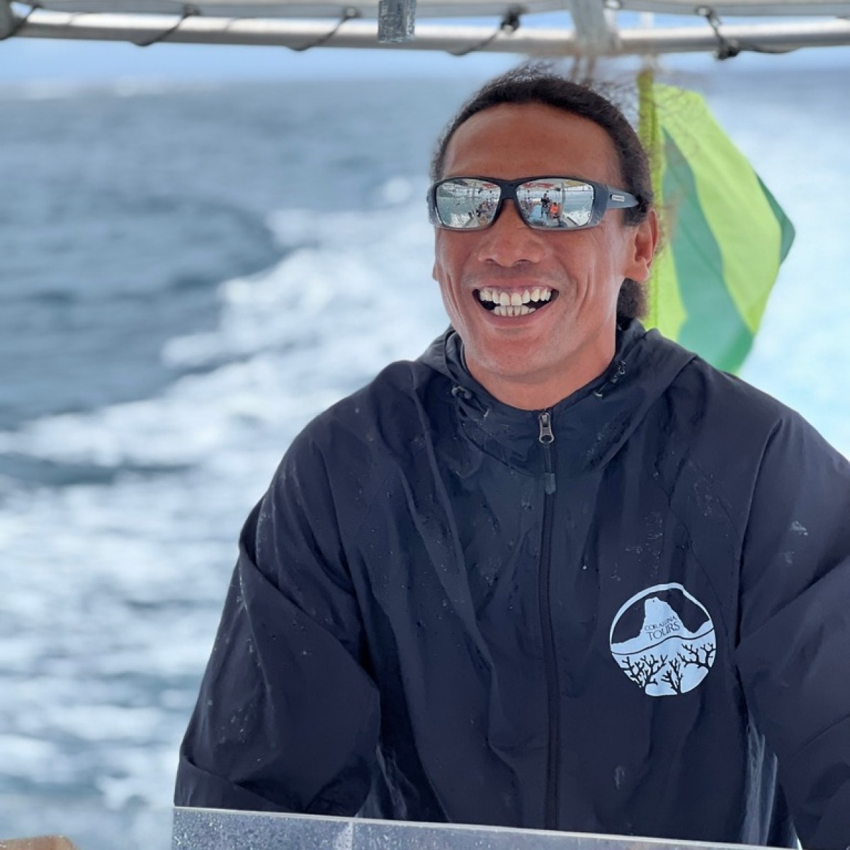Person smiling while steering a boat, wearing sunglasses and a jacket.
