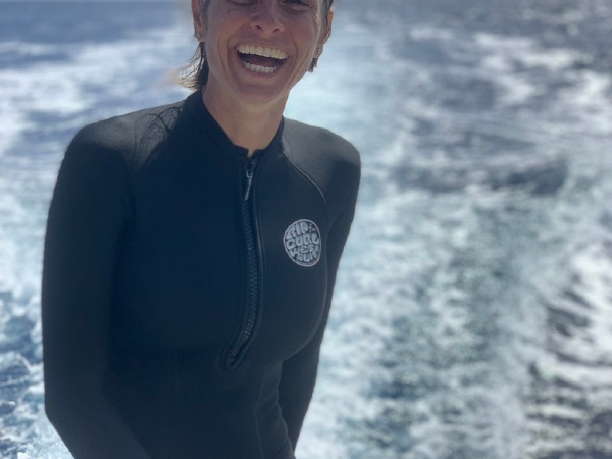 Person in wetsuit smiling on boat with ocean backdrop.